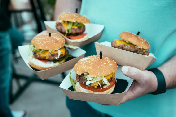 Four freshly made cheeseburgers in kraft-paper trays, held out on a tray by a member of quick-service restaurant staff.