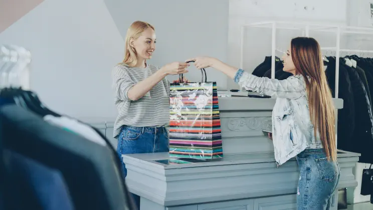 A retail store checkout: an assistant hands a customer their shopping bag